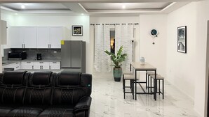 A modern living room with a black leather sofa, a dining area with a table and chairs, a kitchen with white cabinets and appliances, and a large potted plant.