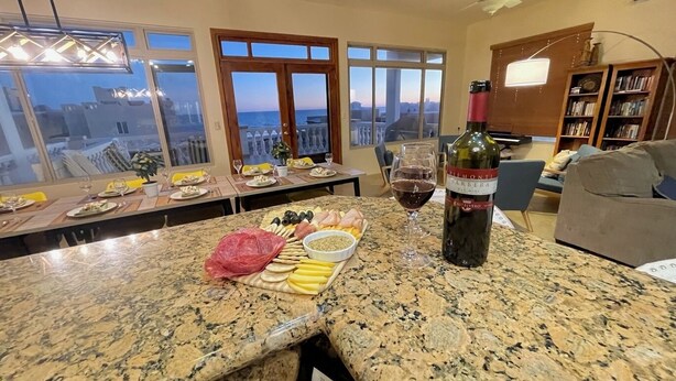 Un coin repas avec une table dressée pour le dîner, une bouteille de vin et une vue sur la mer par de grandes fenêtres.