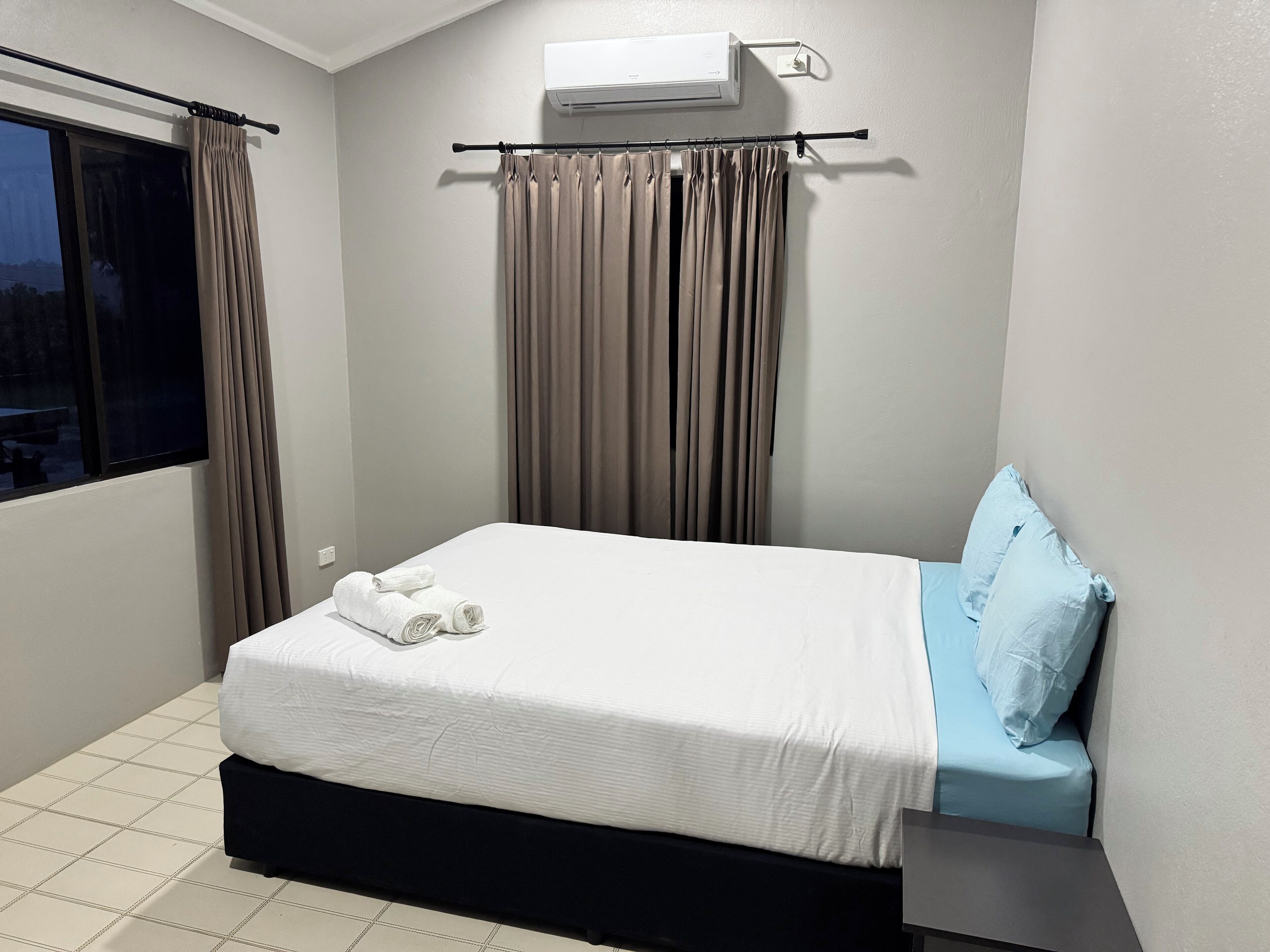 A bedroom with a bed, a window, curtains, an air conditioner, and a view of the outside.