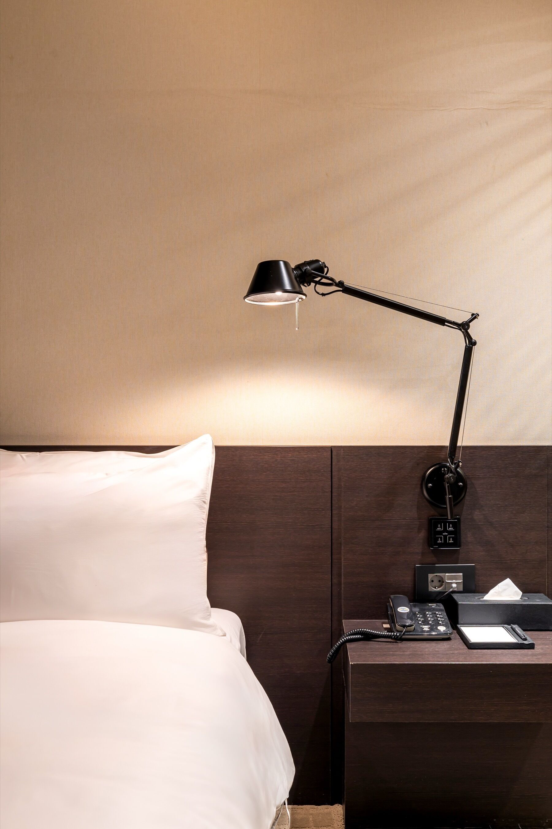 A neatly made bed with white linens, a wooden headboard, a bedside table with a lamp, a telephone, and a tissue box.