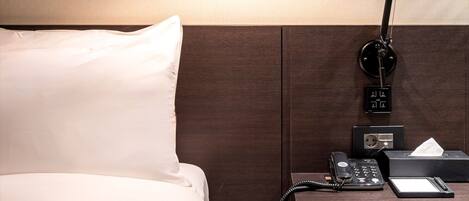A neatly made bed with white linens, a wooden headboard, a bedside table with a lamp, a telephone, and a tissue box.