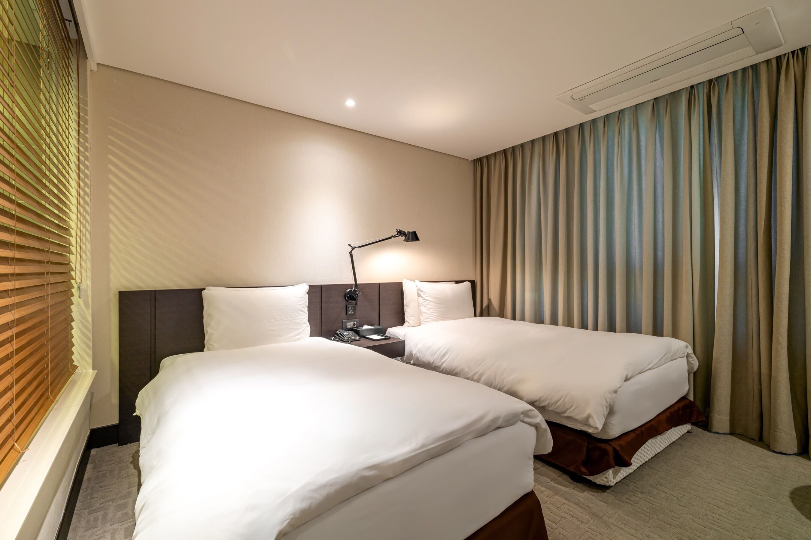 Habitación de hotel con dos camas, una mesita de noche, una lámpara y una ventana con persianas.
