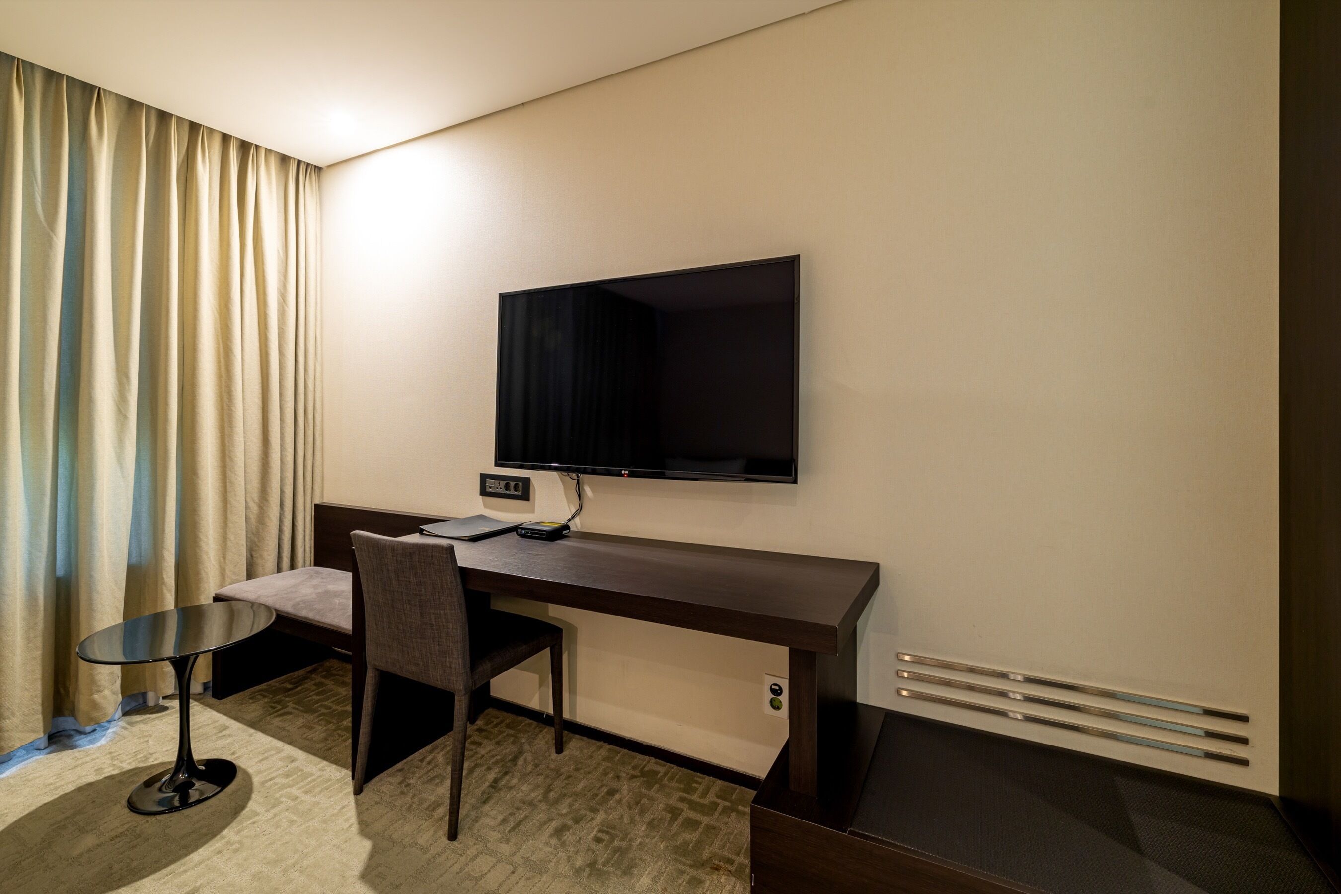 Une chambre d’hôtel avec un bureau en bois foncé, un téléviseur à écran plat fixé au mur, une petite table ronde, une chaise et un lit avec une tête de lit.