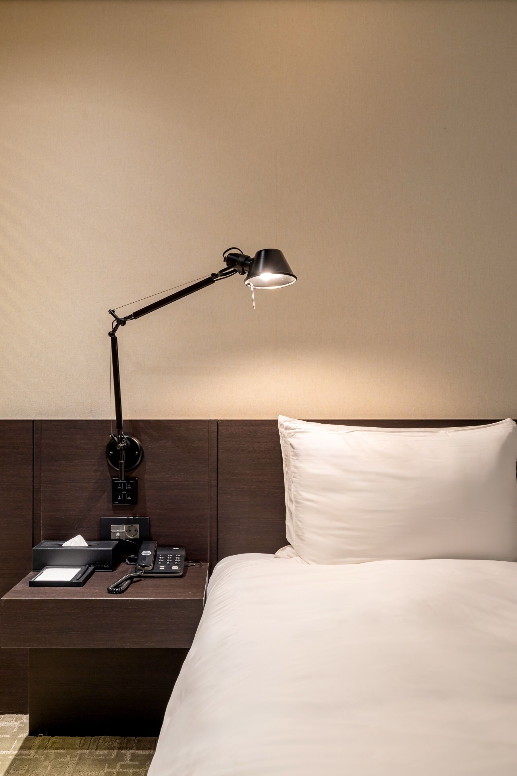 Una cama bien hecha con un cojín blanco y un cabecero de madera oscura. Una lámpara de escritorio negra está sobre la mesita de noche.