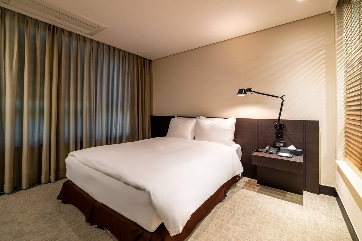 A hotel room with a large bed, a bedside table with a lamp, and a window with blinds.