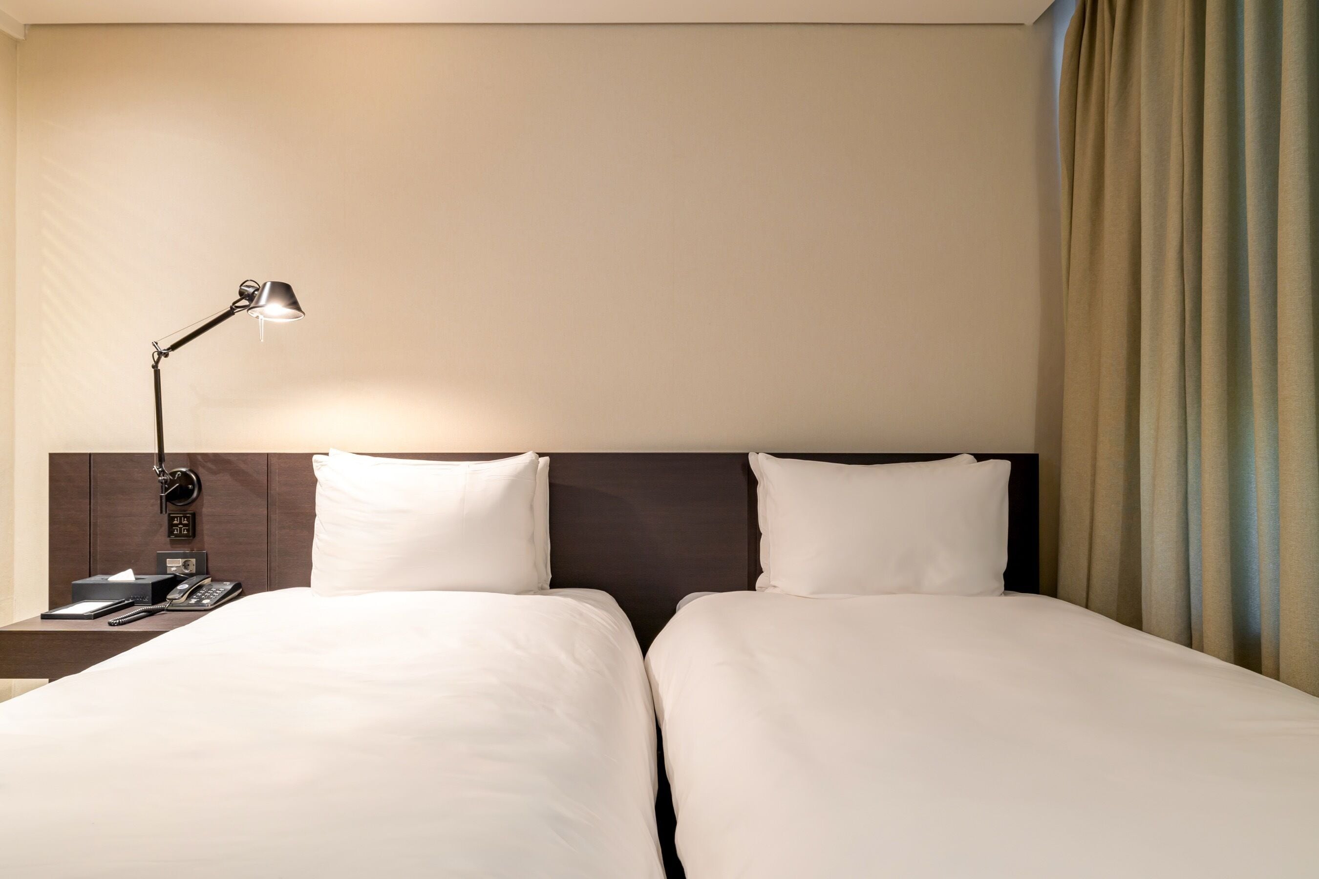 A hotel room with two beds, a nightstand with a lamp, a phone, and a window with curtains.