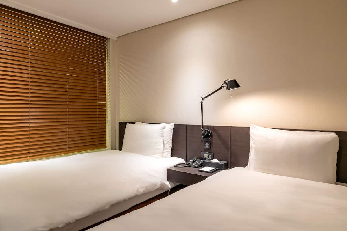 A hotel room with two beds, a bedside table with a lamp, a phone, and a small box.