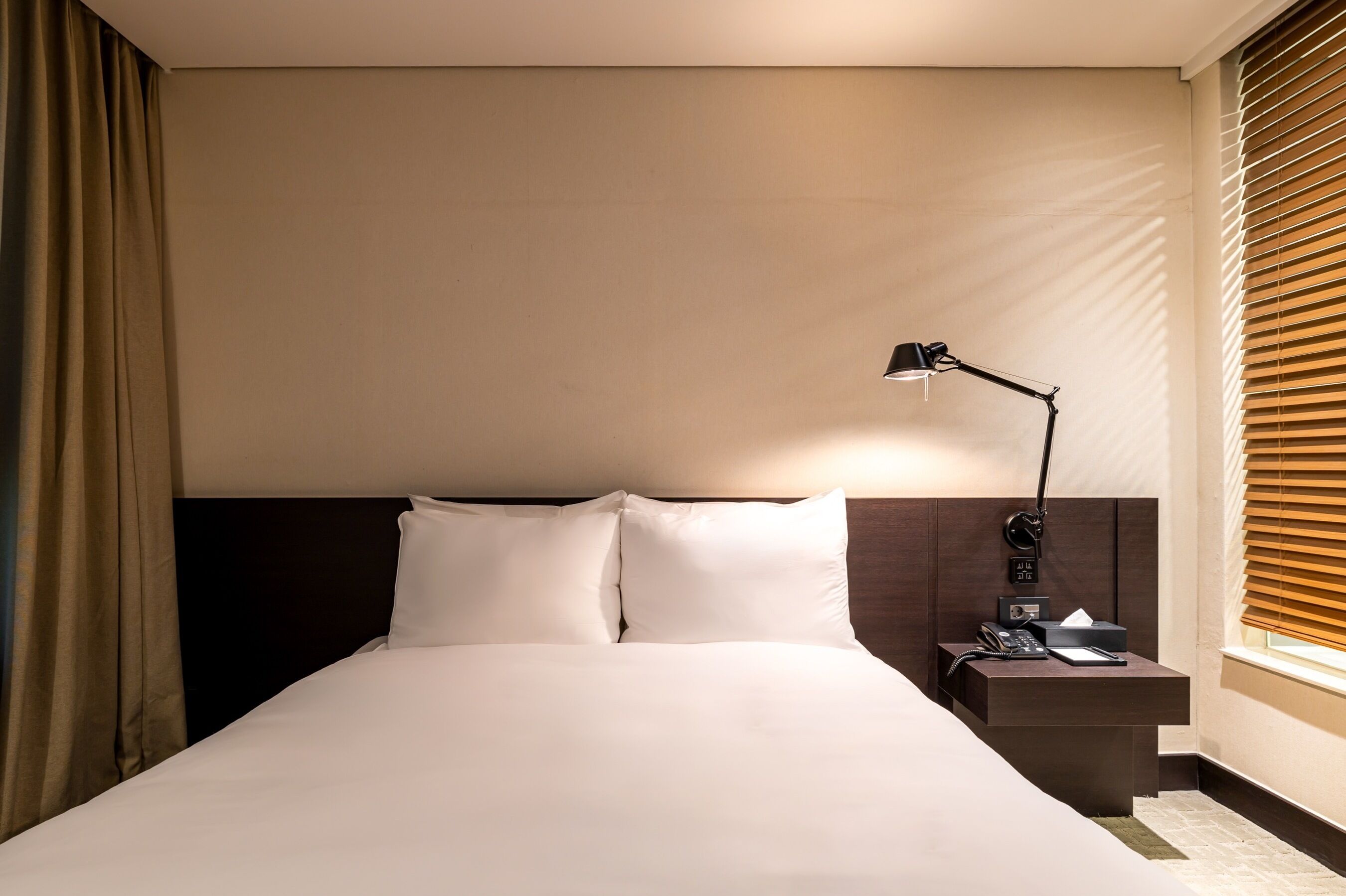 A hotel room with a neatly made bed, a bedside table with a lamp, and a window with blinds.