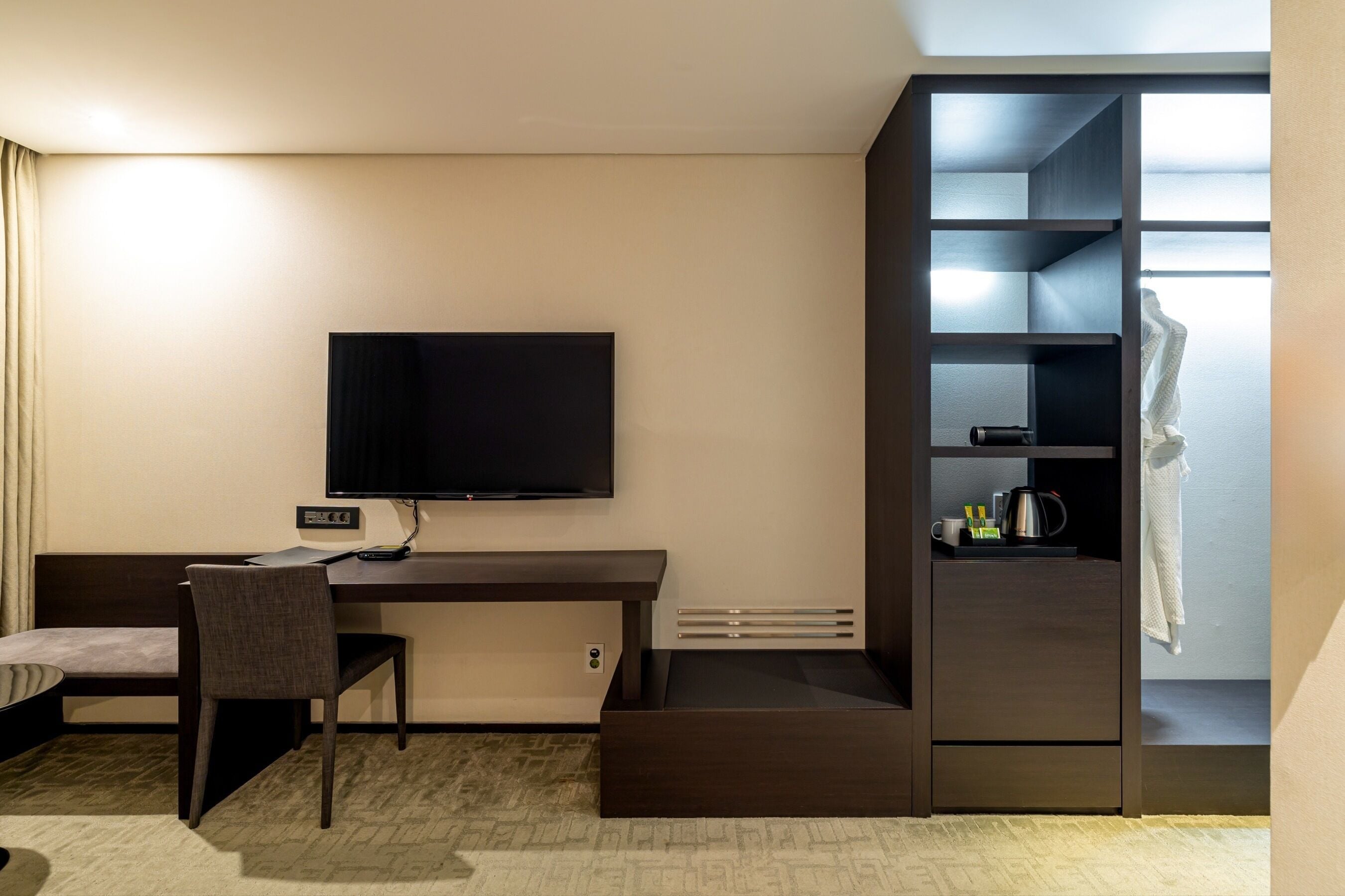 Et moderne hotelværelse med et vægmonteret fladskærms-tv, et skrivebord med stol, indbygget garderobeskab og et lille siddeområde.