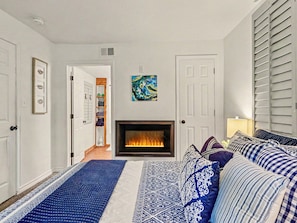 Een woonkamer met een open haard, een bank met gekleurde kussens, een deur naar een andere kamer en een kunstwerk aan de muur.