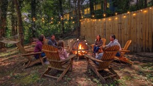 Eine Gruppe von Menschen sitzt um eine Feuerstelle im Freien, mit Holzsesseln und einer Hängematte.