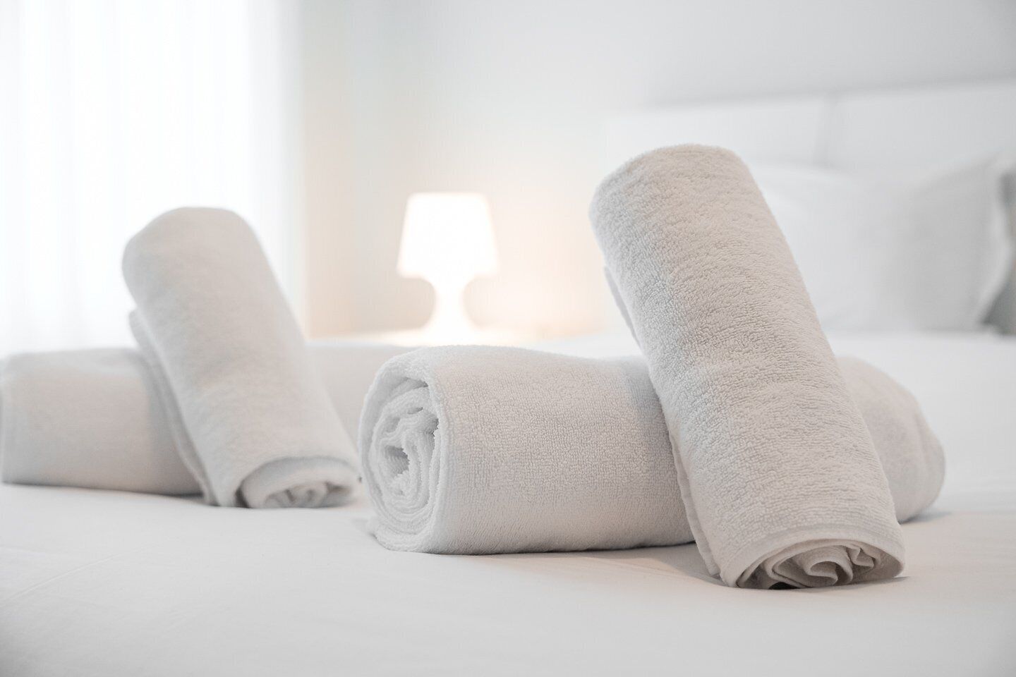 Ein ordentlich bezogenes Bett mit aufgerollten weißen Handtüchern und einer Lampe im Hintergrund.