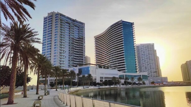 A waterfront view with modern high-rise buildings, a pedestrian walkway, and palm trees.