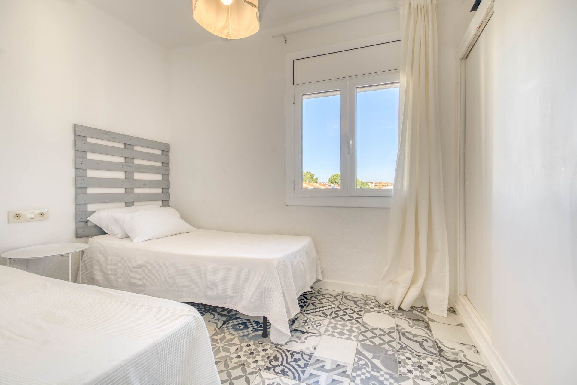 A small, bright room with two beds, a window with curtains, and patterned floor tiles.