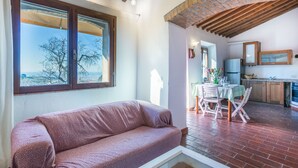 Un salon confortable avec un canapé, un coin repas avec une table et des chaises, une cuisine équipée de placards et d’un réfrigérateur, et une grande fenêtre offrant une vue sur les arbres et les bâtiments.