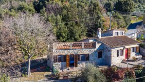 Une maison en pierre dotée d’un balcon, entourée d’arbres et de collines.