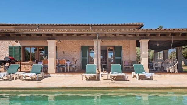 A pool area with clear water, a covered patio with outdoor seating, and a stone building with green shutters. - Charming finca with large pool and garden in Santanyi for up to 8 persons (Santanyi)