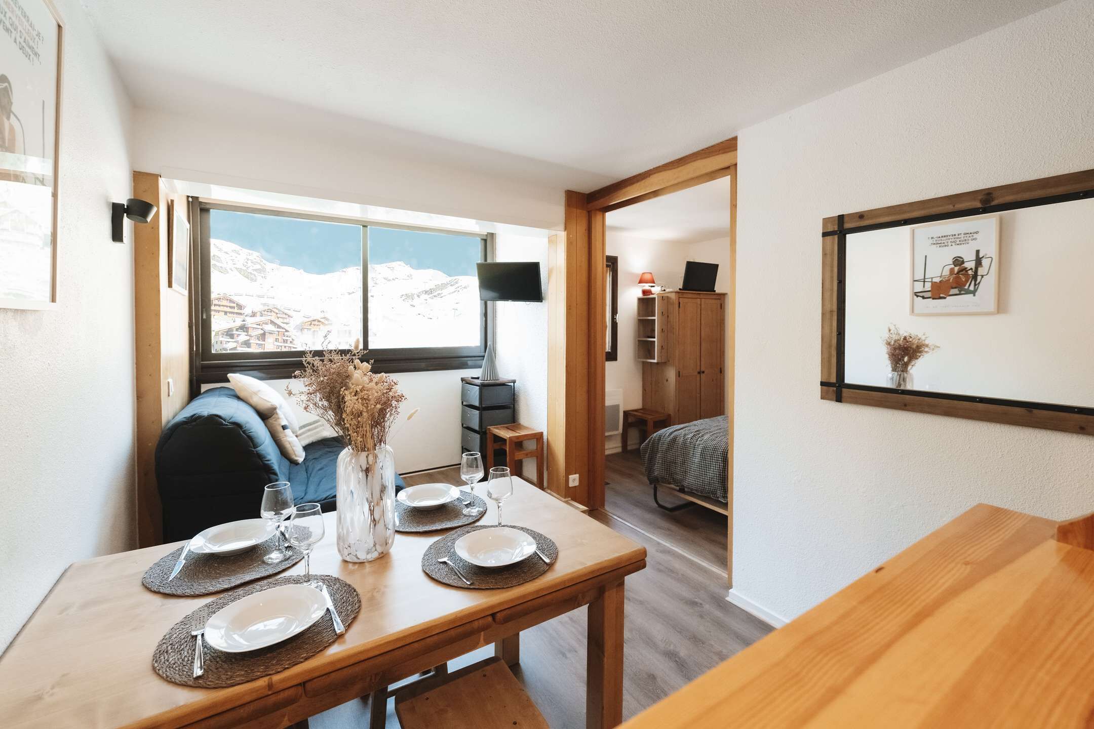 Un'area pranzo con un tavolo apparecchiato per quattro persone, una finestra vista montagne innevate e un armadio in legno sullo sfondo.