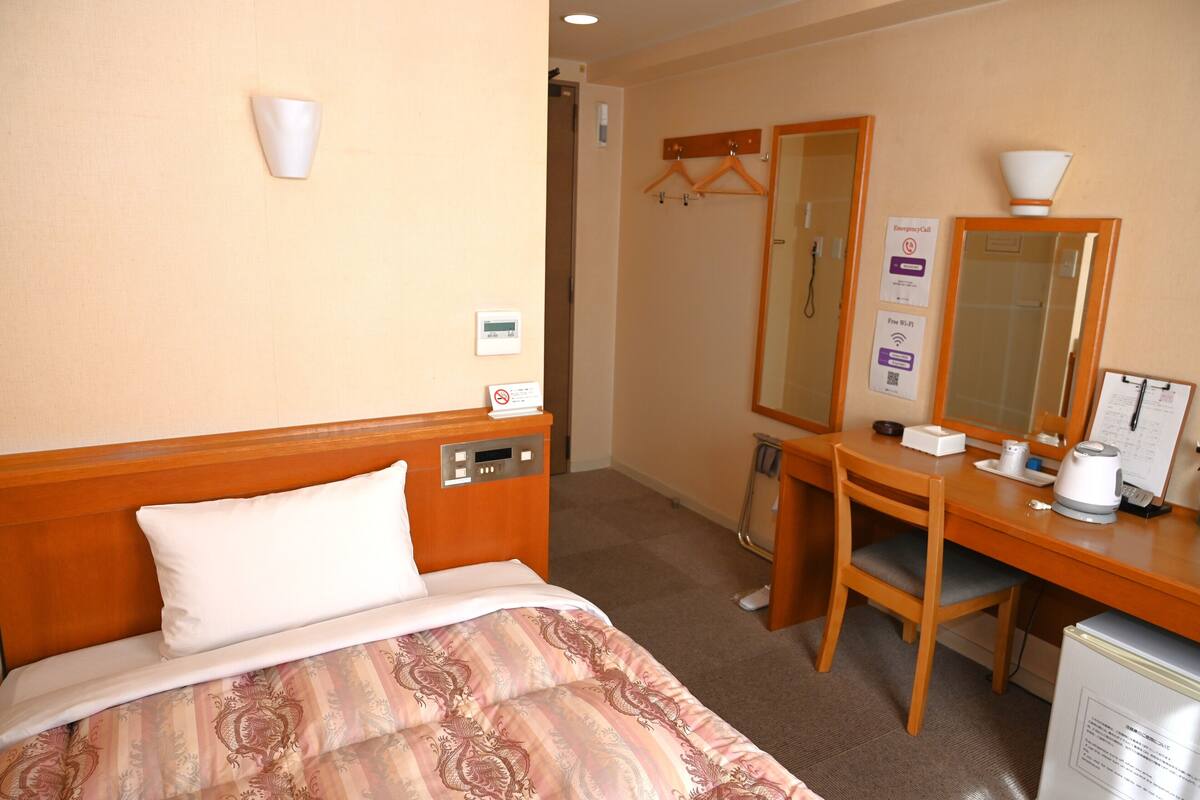 A hotel room with a bed, bedside table, a desk with a chair, a mirror, and a small refrigerator.