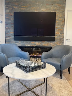Una sala de estar moderna con chimenea de piedra, un televisor de pantalla plana montado encima, dos sillones grises y un tablero de ajedrez sobre una mesa de centro redonda.