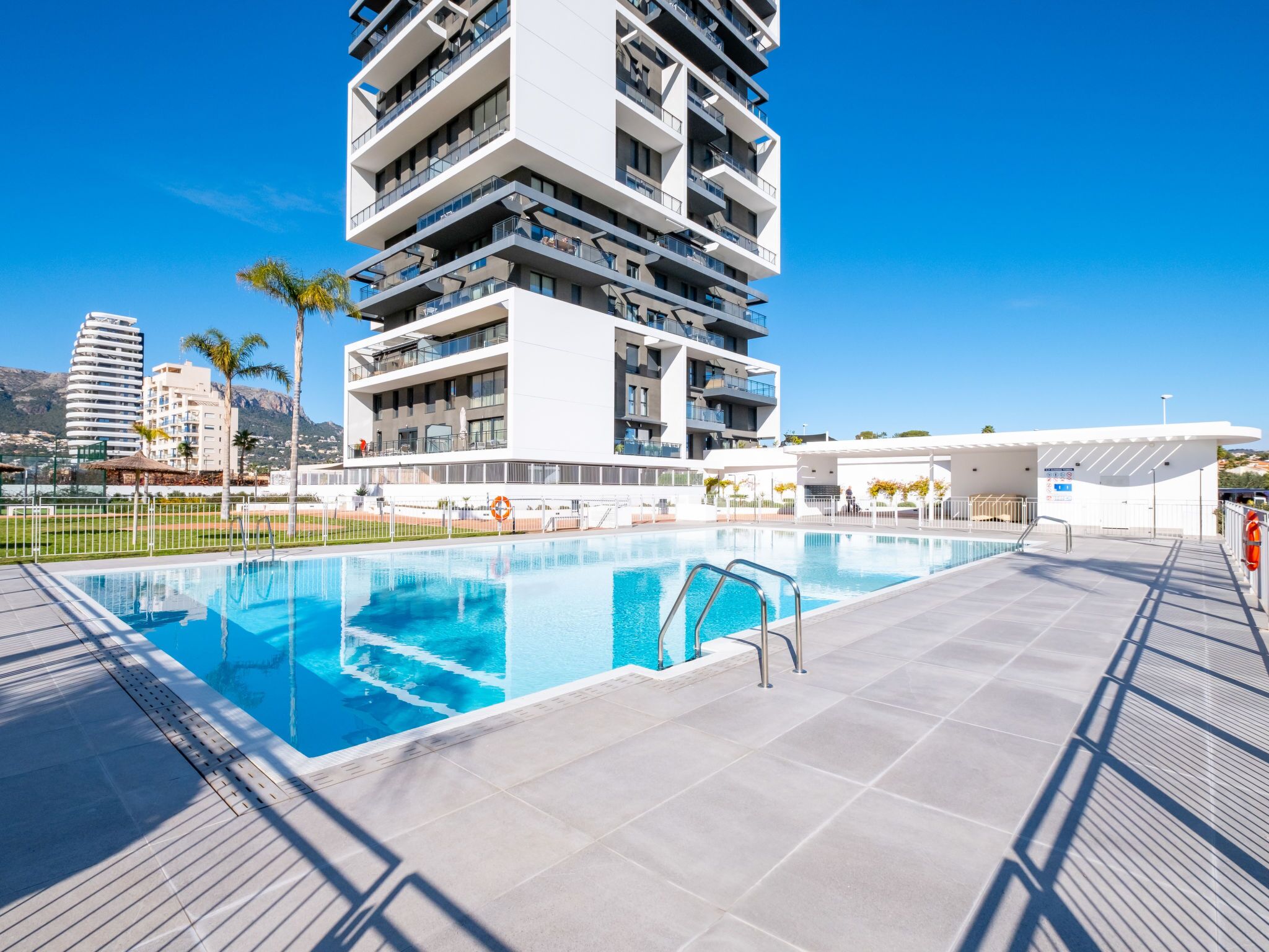 A modern building with a rooftop pool, surrounded by a fence and palm trees.