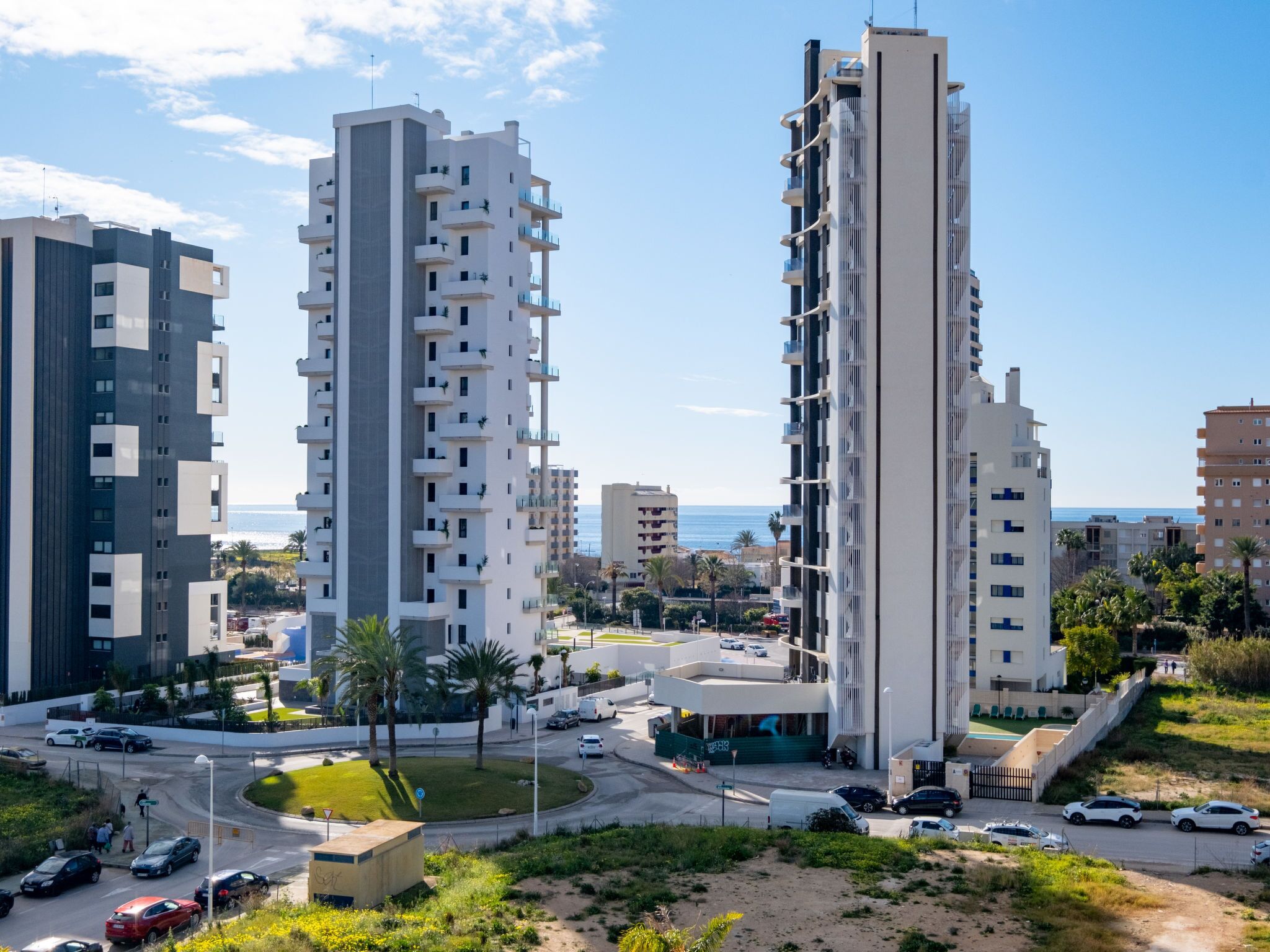 A modern high-rise building with multiple balconies, surrounded by other high-rise buildings and a coastal view.