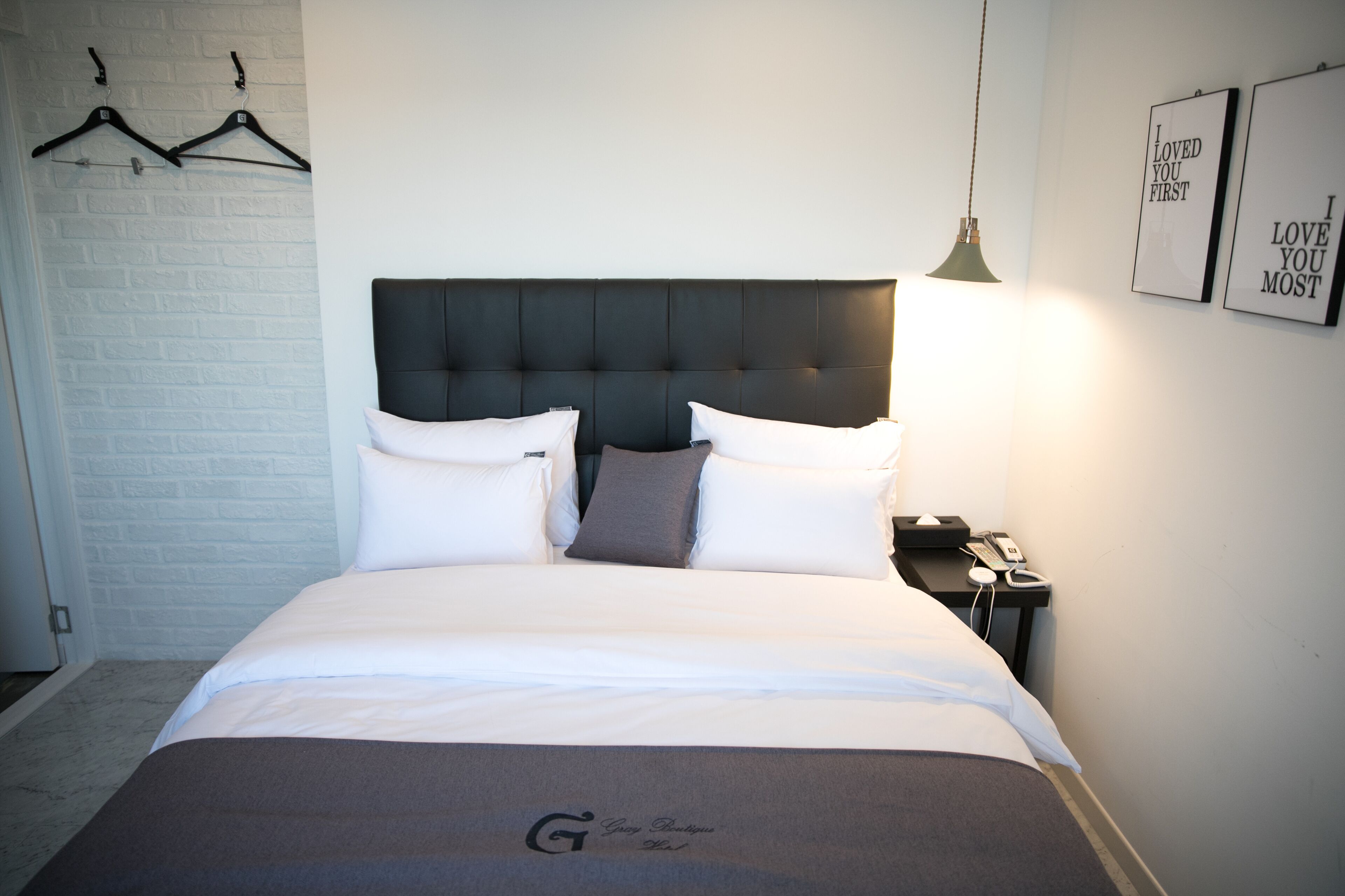 A neatly made bed with a dark headboard, white pillows, and a grey throw. There is a bedside table with a lamp and a phone. The wall has a white brick pattern and two framed pictures with text.