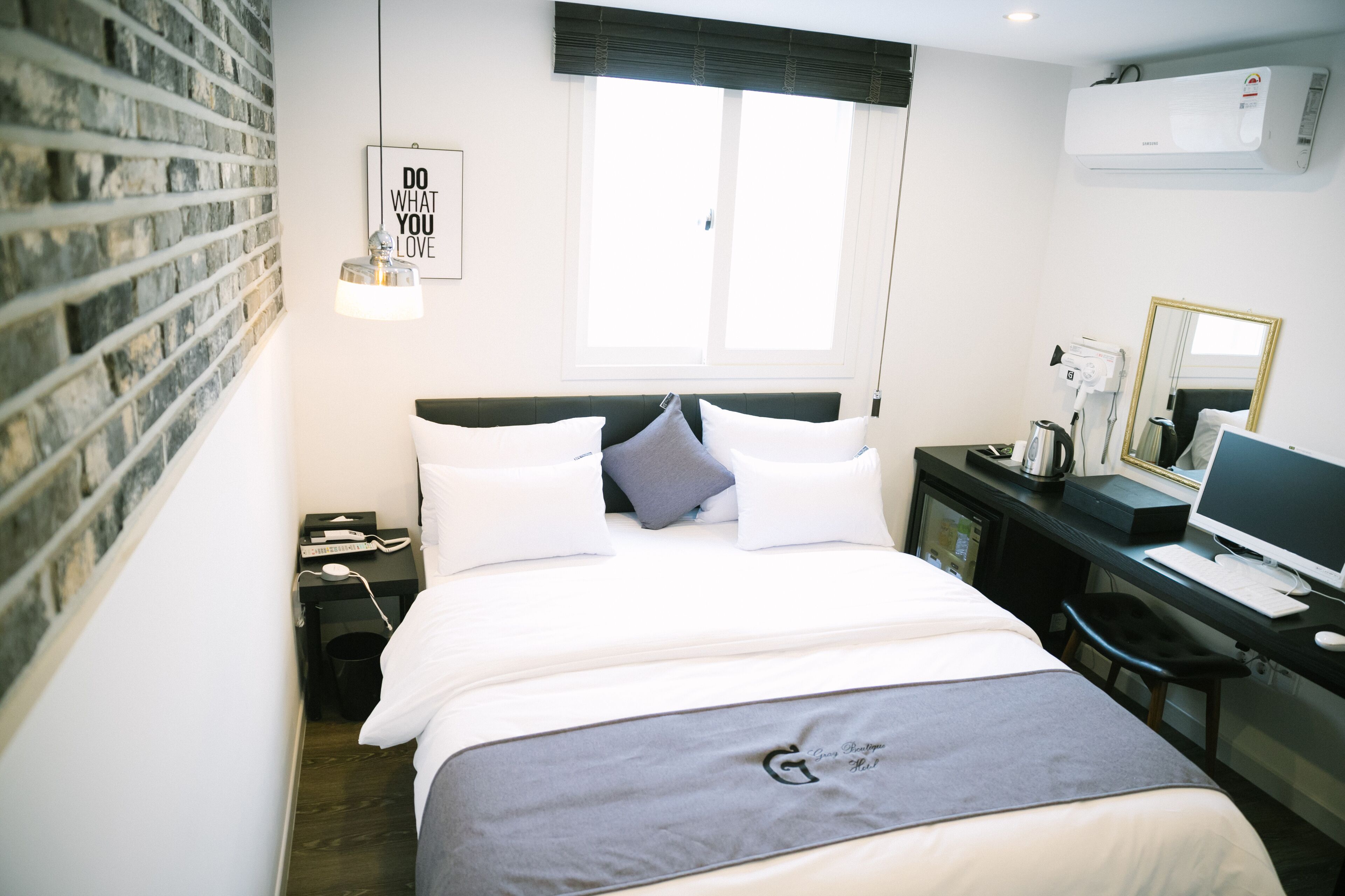 Une chambre d’hôtel avec un mur en briques, un lit avec une literie blanche, un bureau avec un ordinateur et une fenêtre avec des stores.
