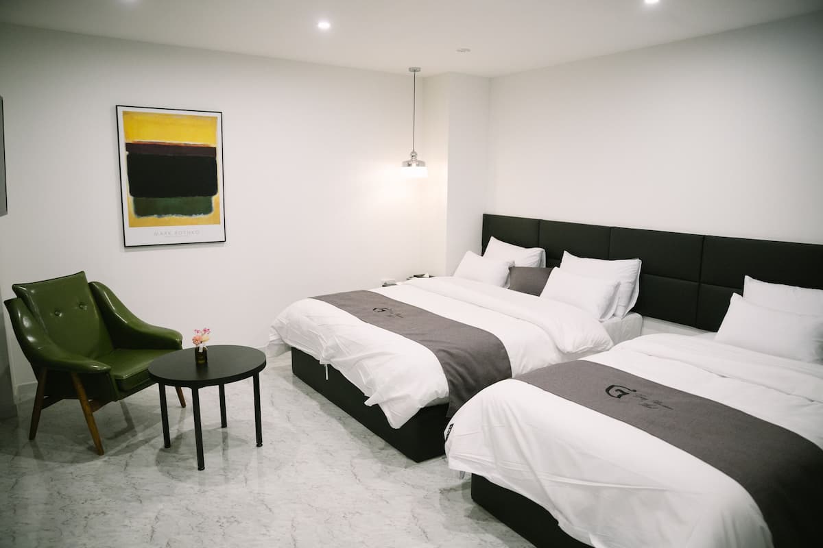 A hotel room with two beds, a green chair, a small table, and a painting on the wall.