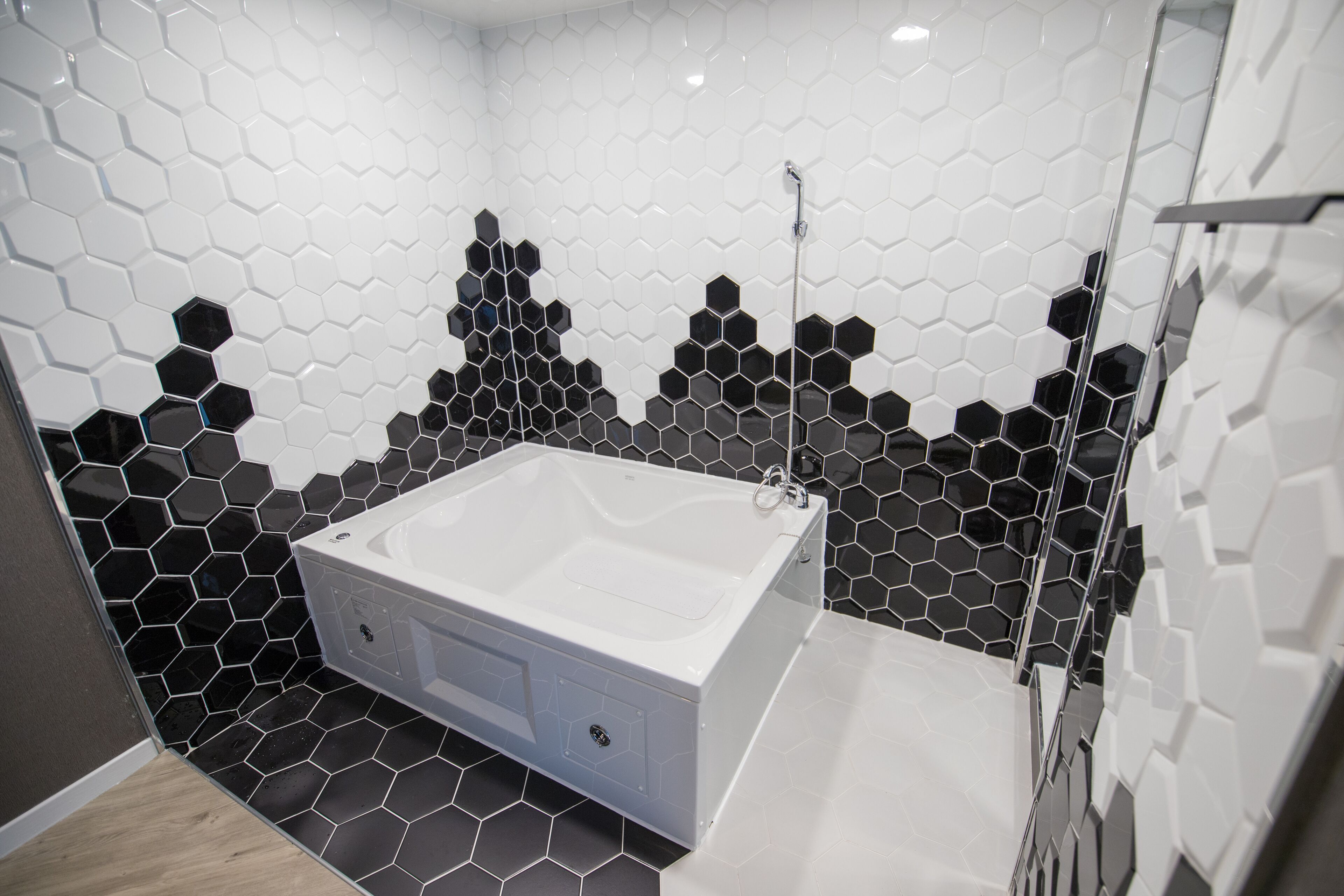 A modern bathroom with a large white bathtub, black and white hexagonal tile walls, and a wooden floor.