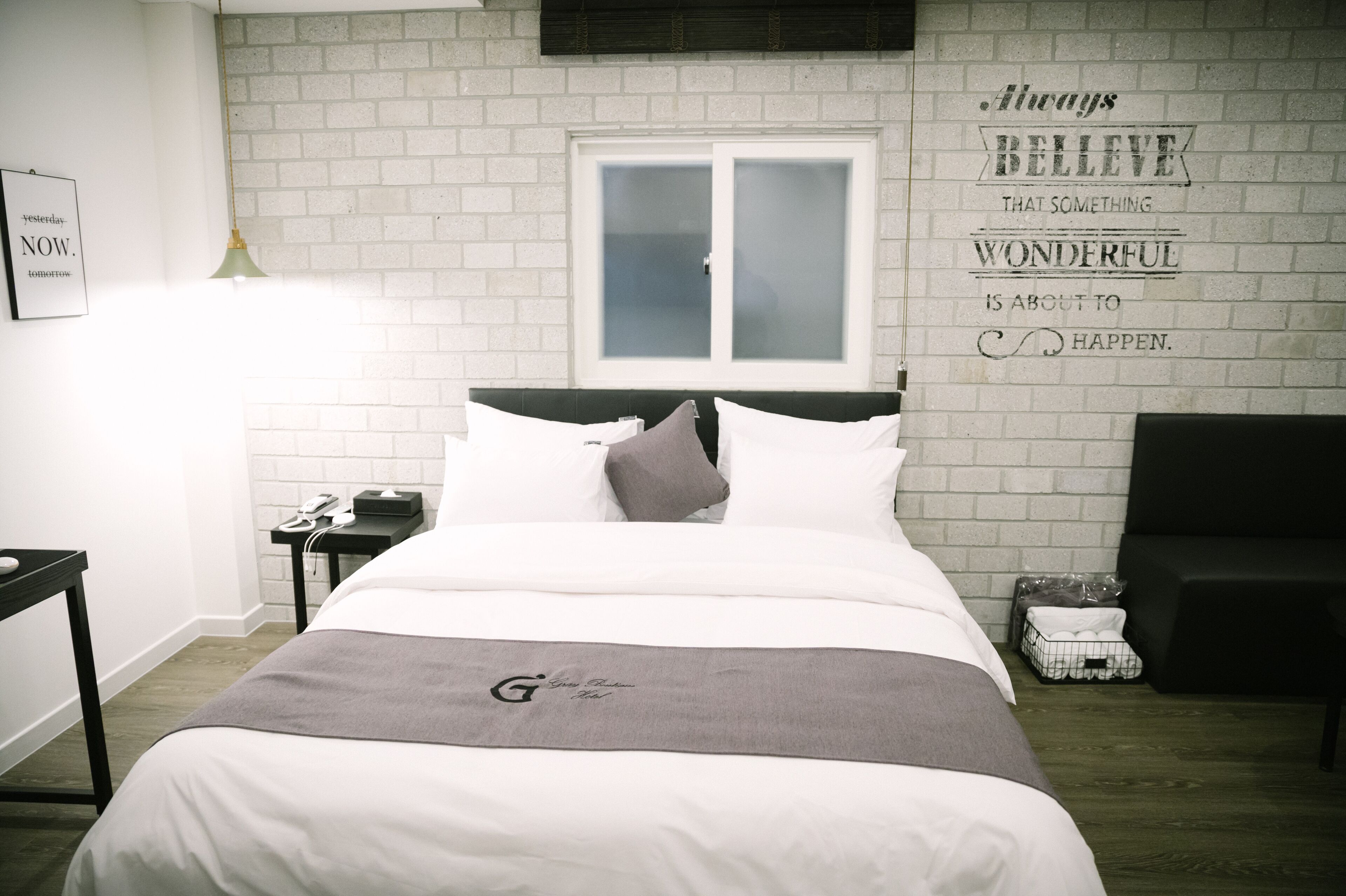 Habitación de hotel ordenada con una cama, una mesita de noche, una lámpara, un ventanal y una frase motivadora en la pared.