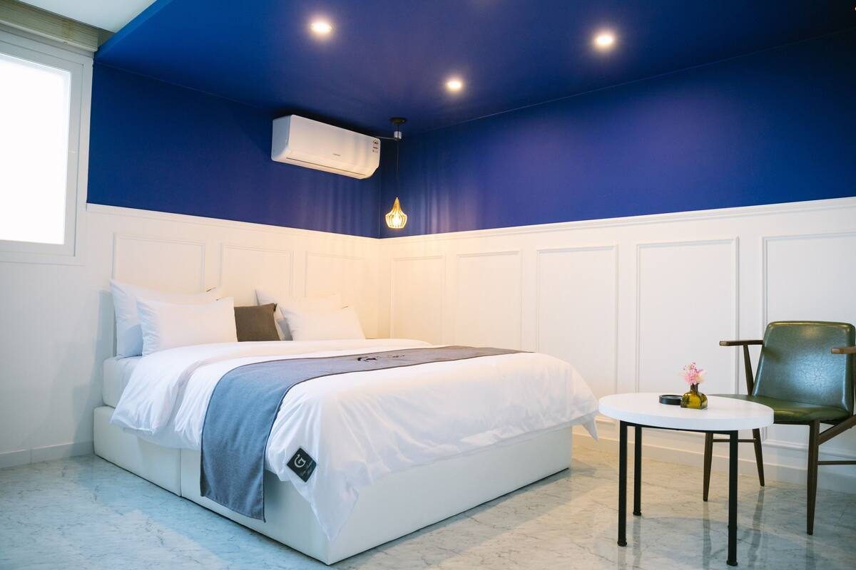 A bedroom with a bed, a chair, a small table, and an air conditioning unit.