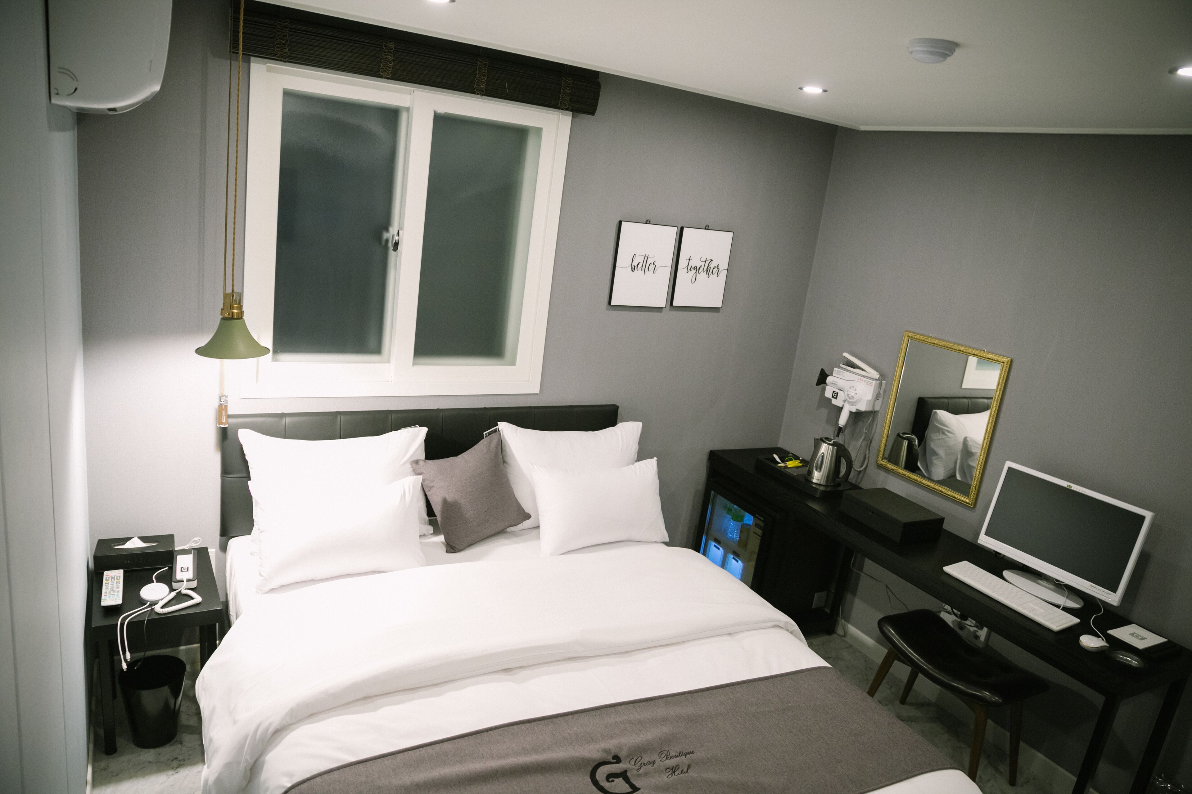 A hotel room with a bed, bedside table, desk, chair, computer, and a mirror.