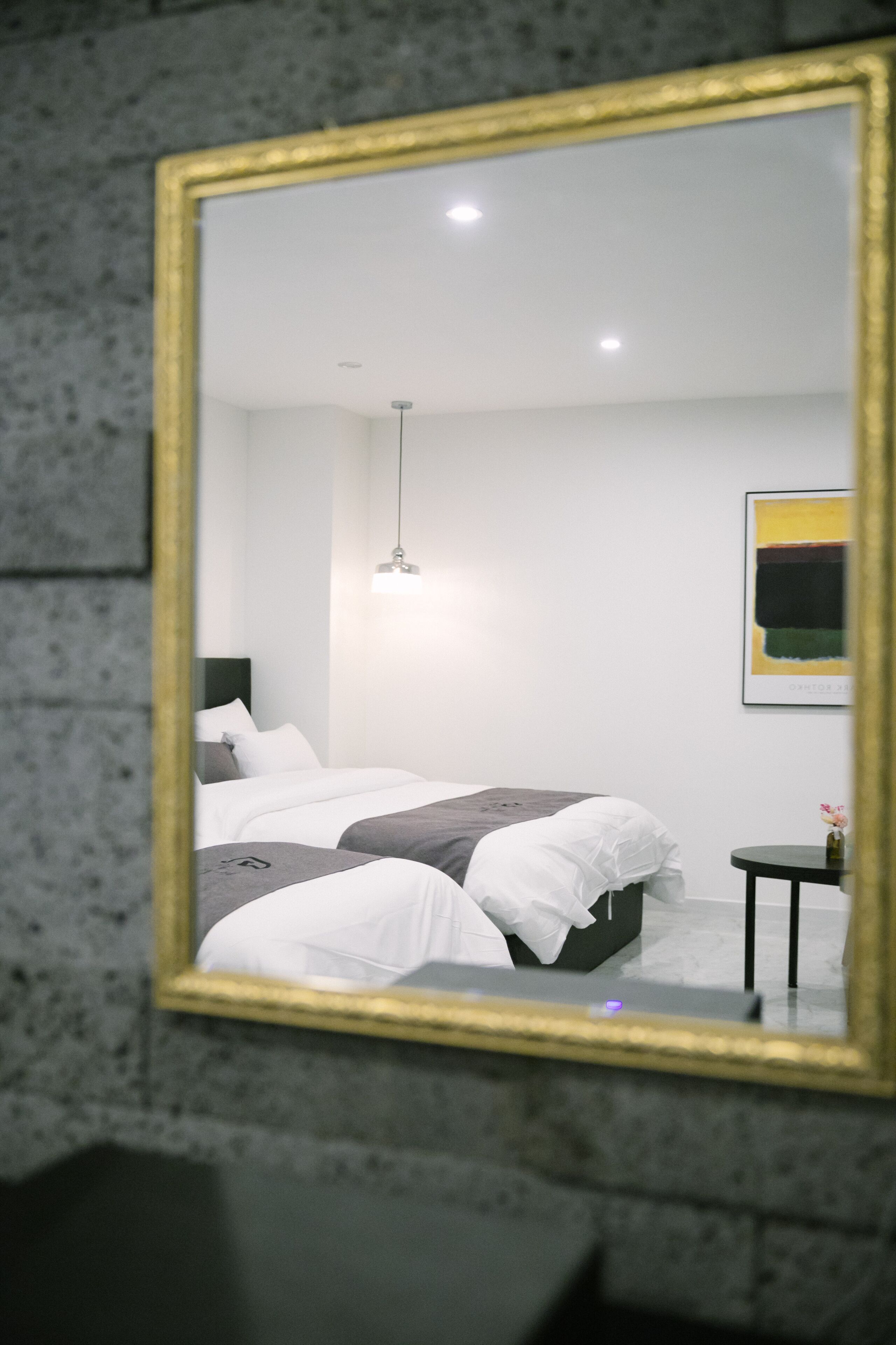 Quarto moderno com um espelho grande, cama com roupa de cama branca e cinza, mesa de cabeceira pequena e luminária pendente.