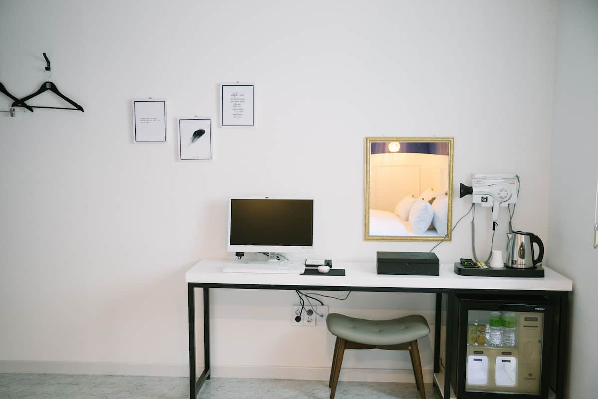 A modern hotel room with a white desk, a computer, a mirror, a hairdryer, a kettle, and a minibar.