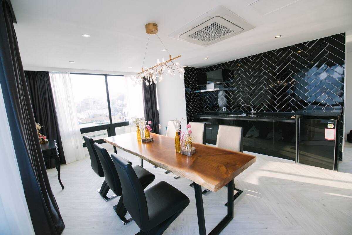 A modern dining area with a wooden table and black chairs, a black herringbone backsplash in the kitchen, and a large window with curtains.