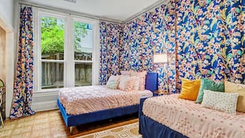 A bedroom with two beds, floral wallpaper, a patterned rug, and a window with curtains.
