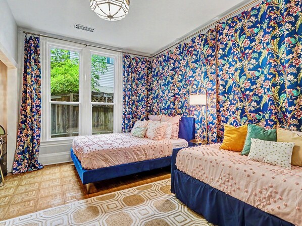 A bedroom with two beds, floral wallpaper, a patterned rug, and a window with curtains.