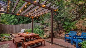 A wooden pergola with string lights, a wooden table with benches, and blue chairs. - Walk to Bathhouse Row! Queen Anne Victorian Cottage (Hot Springs)