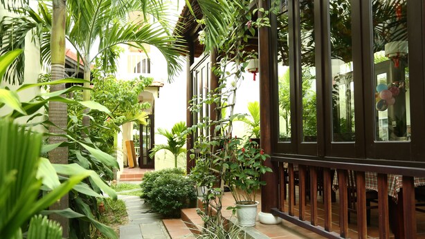 Un étroit sentier bordé d’une végétation luxuriante et de plantes en pot, qui mène à un bâtiment doté d’une balustrade en bois et d’un sol carrelé.