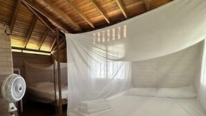 Una habitación con techo de madera, una cama con mosquitero, un ventilador a un lado de la cama y una ventana con luz natural.
