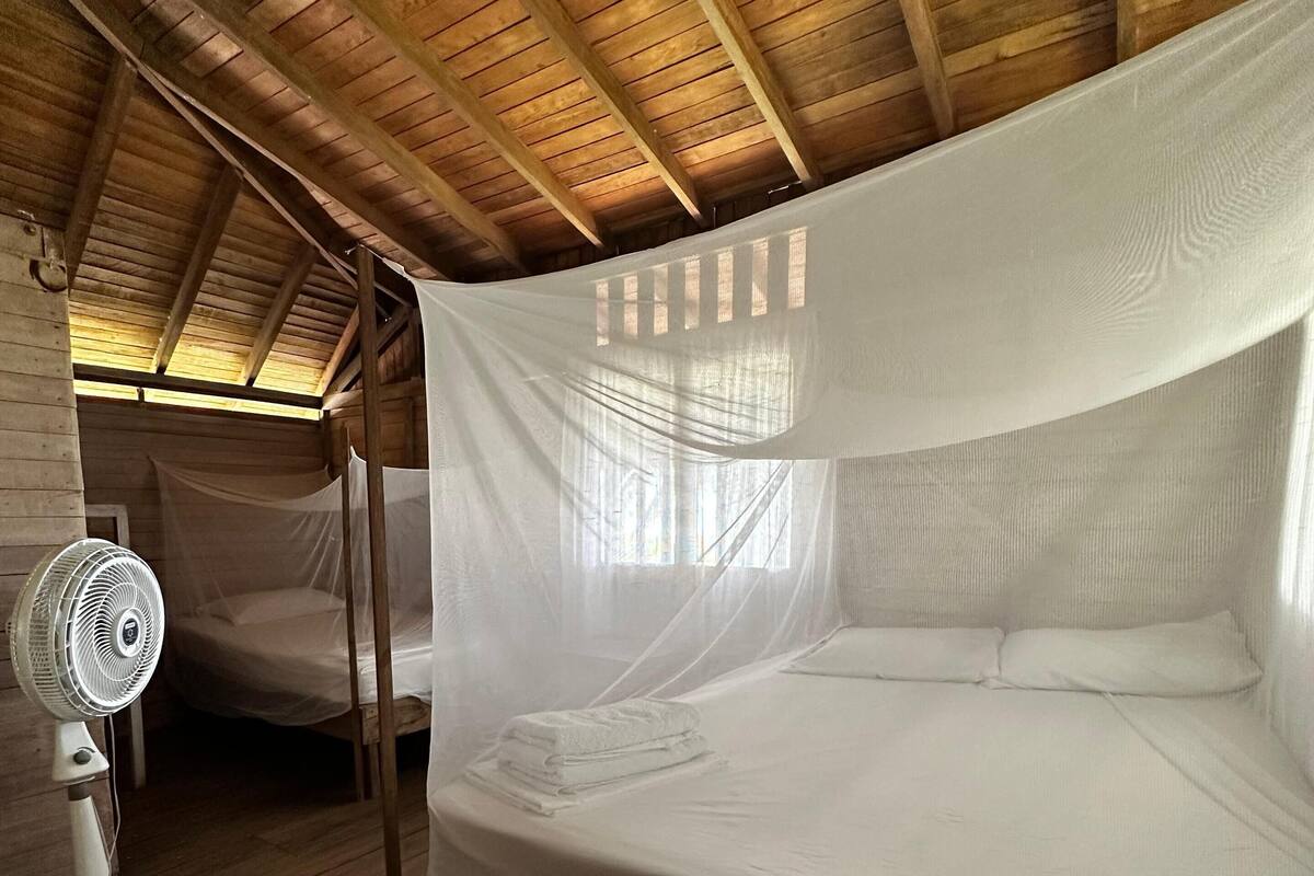 Une chambre avec un plafond en bois, un lit doté d’une moustiquaire, un ventilateur de chevet et une fenêtre laissant entrer la lumière naturelle.