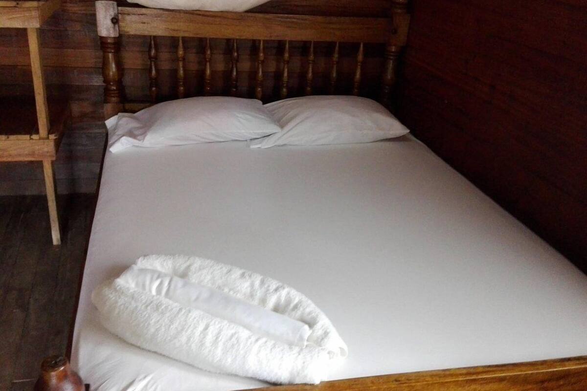 Un lit simple avec du linge de lit blanc et un oreiller blanc, dans une pièce aux murs et à la tête de lit en bois.