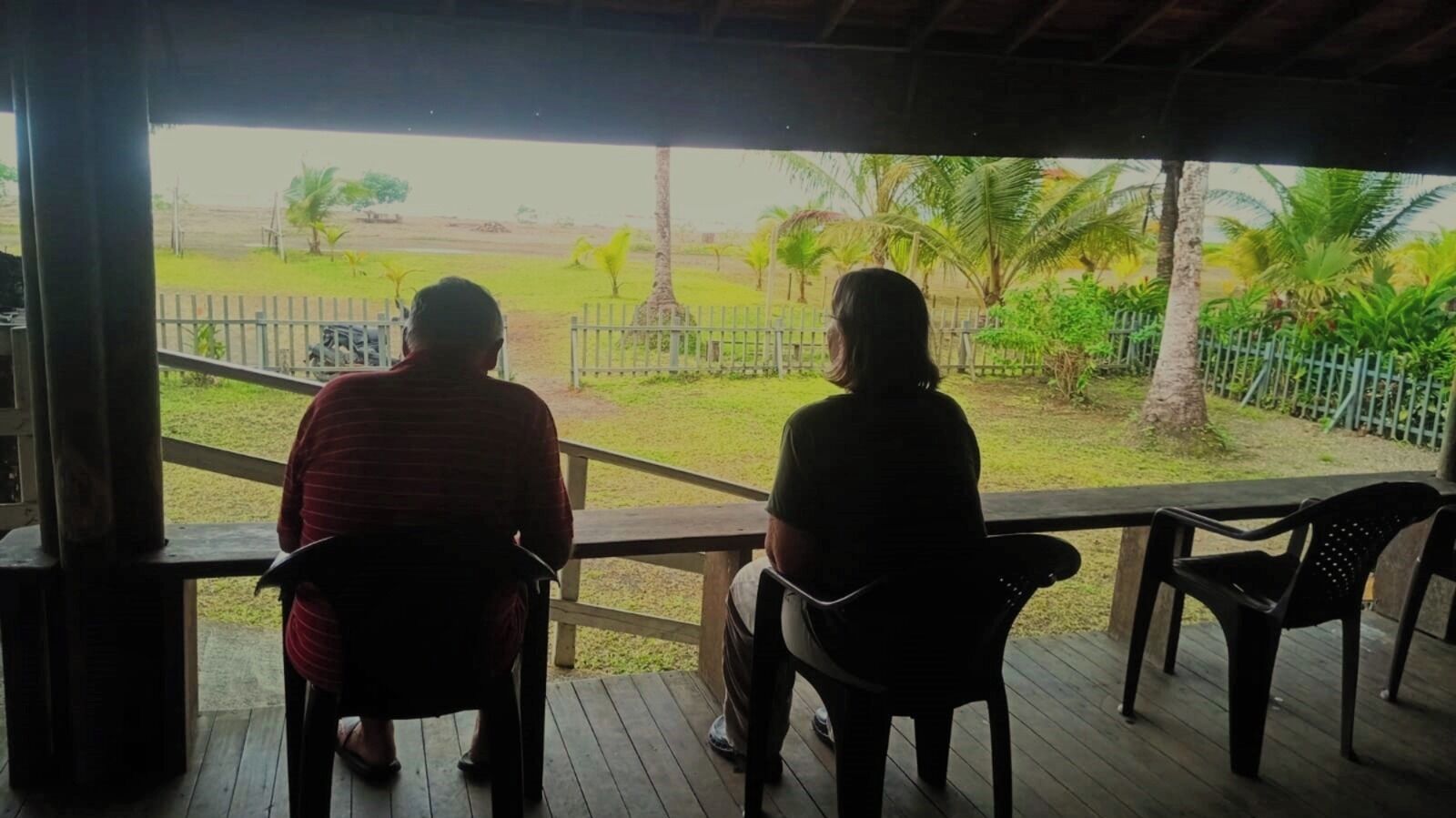 Deux personnes sont assises sur une terrasse en bois, d’où l’on aperçoit une zone délimitée par une clôture et des palmiers.