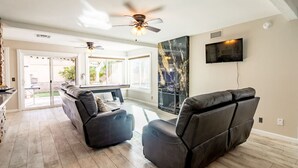A living room with a fireplace, a ceiling fan, and a flat-screen TV mounted on the wall. - Night at the Roxford 4b 3ba Pool (Las Vegas)