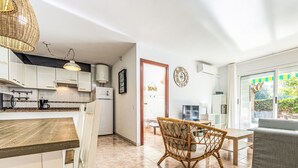 A modern kitchen with white cabinets, a built-in oven, and a coffee machine. There is a dining area with a wicker chair and a grey sofa. A clock and a sign reading "Beach" are visible on the wall.