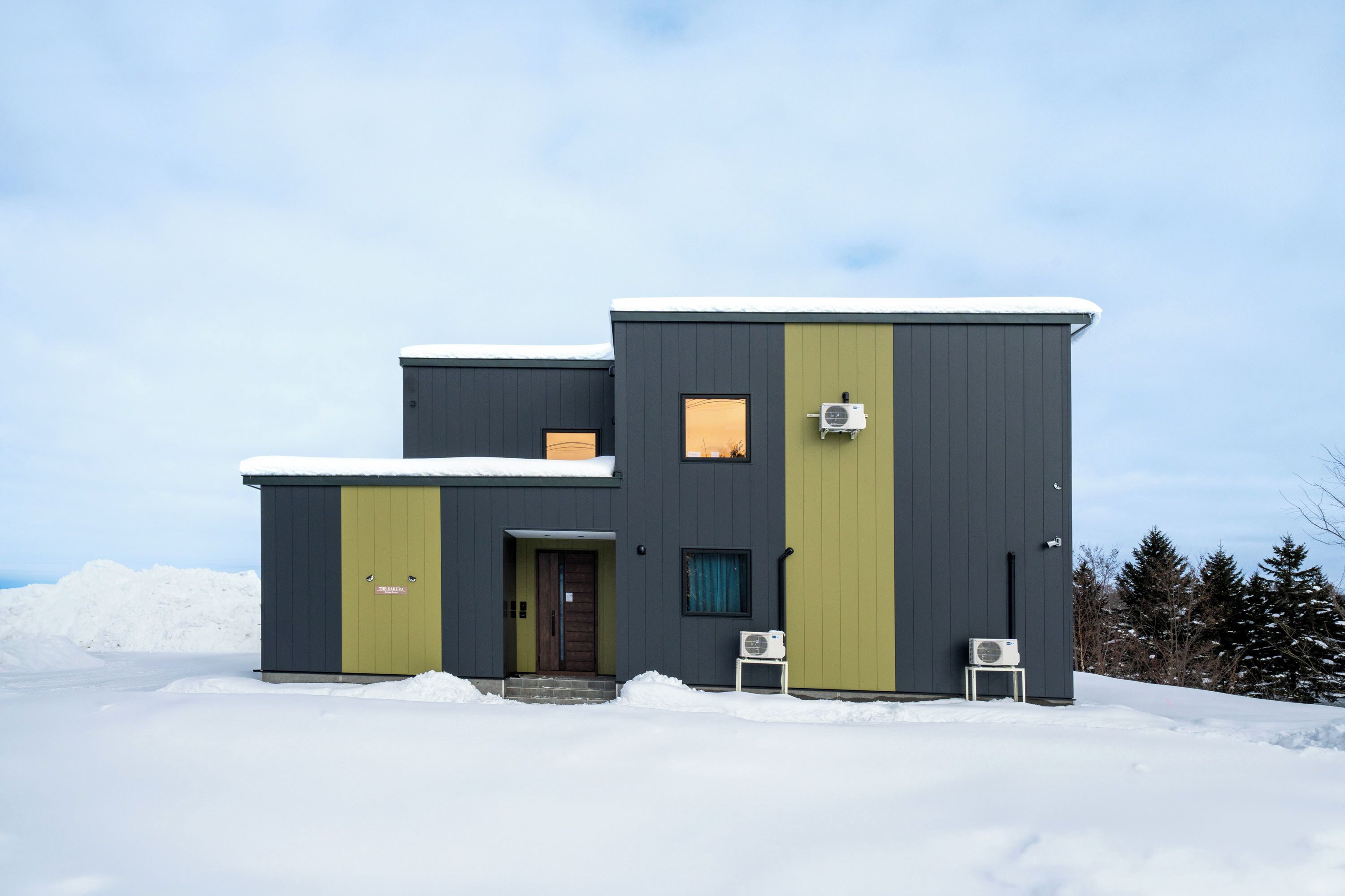 A modern two-story building with a dark exterior and a green door, surrounded by snow.