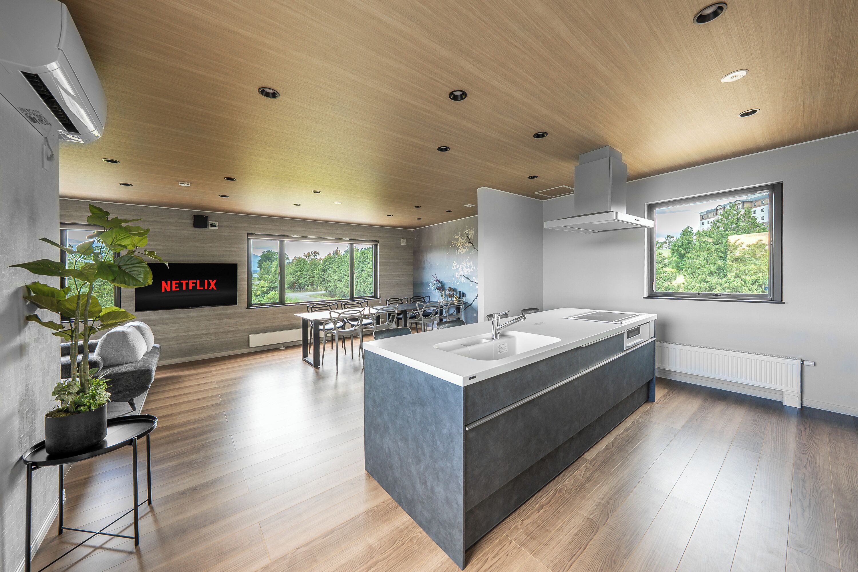A modern kitchen with a central island, a dining area with a table and chairs, and a large window offering a view of greenery.