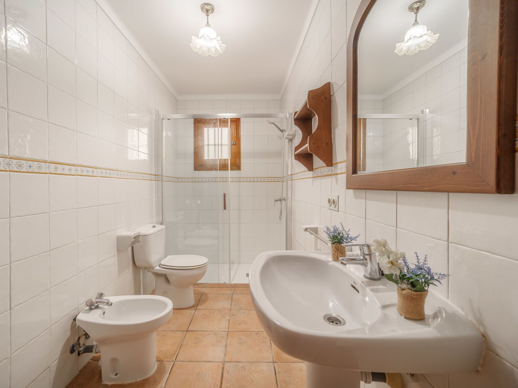 Une salle de bain carrelée de blanc, avec une douche, des toilettes, un bidet et un lavabo surmonté d’un miroir.
