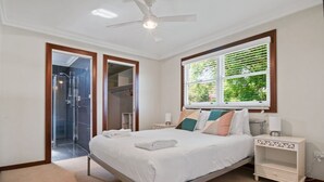 A bedroom with a bed, bedside tables, a window with blinds, a ceiling fan, and a bathroom with a shower.
